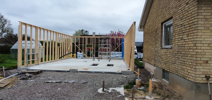 Nybyggd trästomme för hus vid betonggrund nära befintligt tegelhus, byggmaterial och ställning synliga.