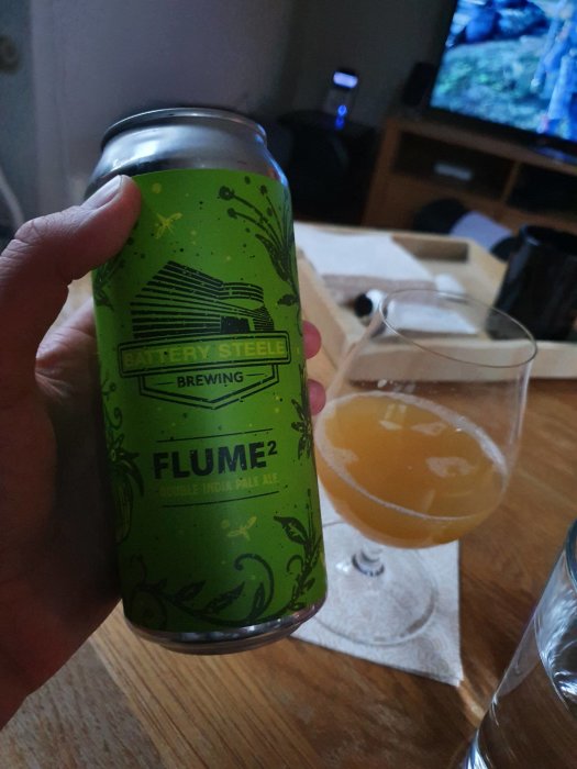 Hand håller en burk Battery Steele Brewing FLUME Double India Pale Ale, med ett glas fyllt med grumligt öl i bakgrunden.