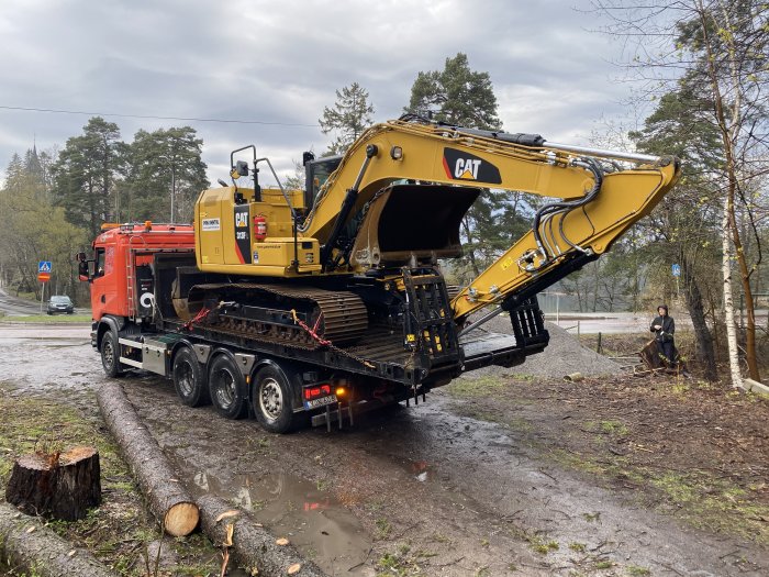 Gul grävmaskin lastas av från en röd lastbil vid en byggarbetsplats, stubbar och schaktmassor syns.