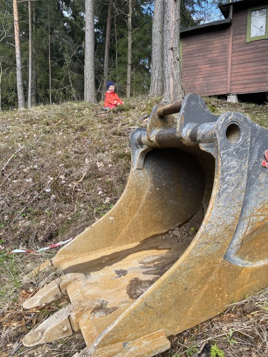 Skopan till en grävmaskin med schaktmassor framför och ett barn i bakgrunden på en tomtrivningsplats.