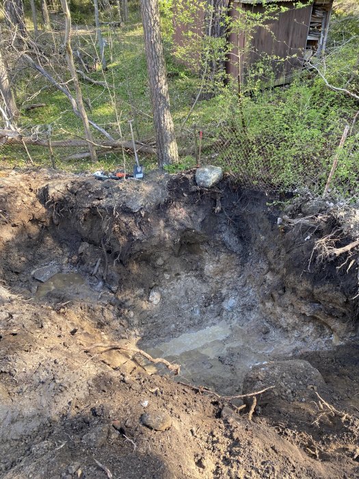 Schaktad grop i skogsmiljö med berggrund och ansamling av vatten, verktyg på kanten.