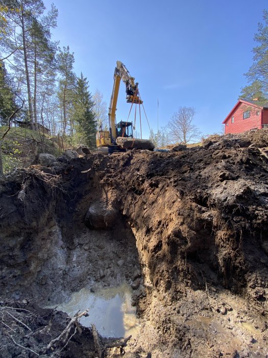 Grävmaskin vid en schaktad grop med synligt grundvatten, omgiven av högar av jord och sten.