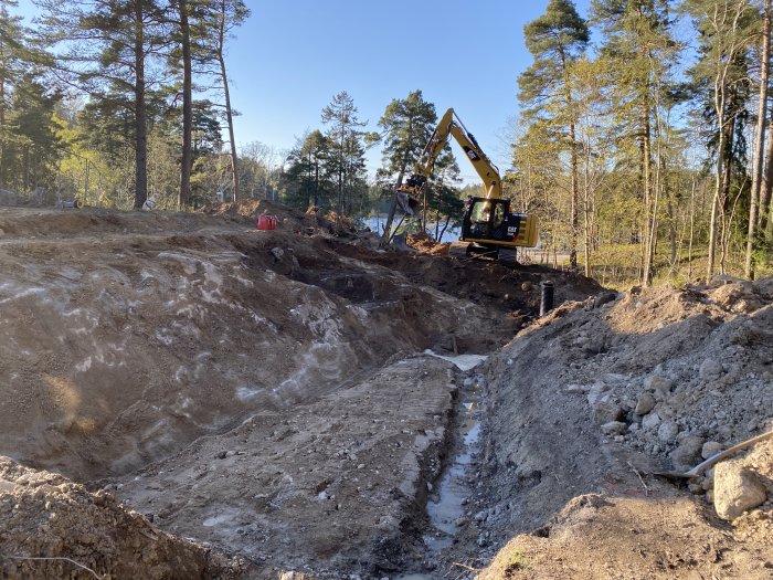 Grävmaskin på en byggarbetsplats med uppschaktade markområden, stenar och berg synliga, bland träd i dagsljus.