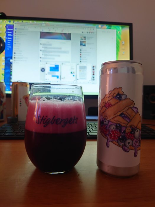 Ett glas fyllt med mörkröd dryck framför en datorskärm, bredvid en burk med färgglad etikett.