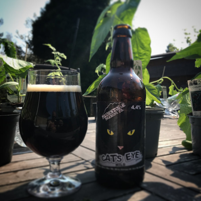 Flaska och glas mörk öl på träbord utomhus nära växter, etikett med kattögon, "Cat's Eye" 4.4%.