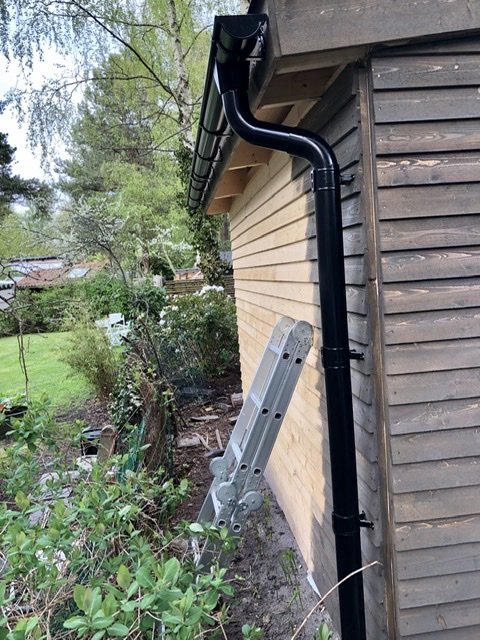 Nyinstallerad svart hängränna med felaktig infästning vid ett trähus bredvid en steg.