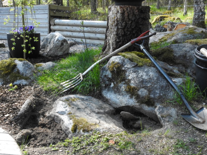 Grävplats med stora stenar, jord och trädgårdsverktyg, samt krukväxter i bakgrunden.