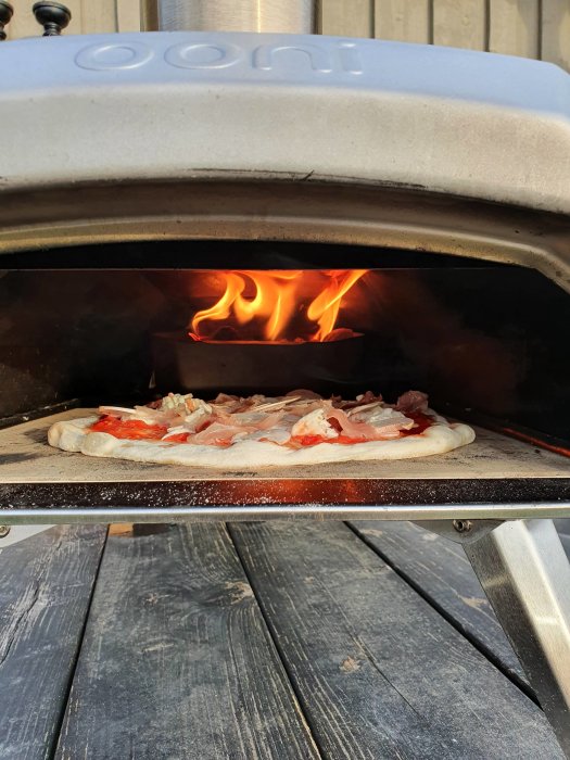 Pizza med skinka som gräddas i en bärbar vedeldad ugn med synliga lågor.