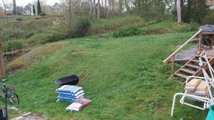 En ojämn gräsmatta med ojämnheter och en backe, några säckar gödsel, en skottkärra och trädgårdsredskap framför en trätrappa.