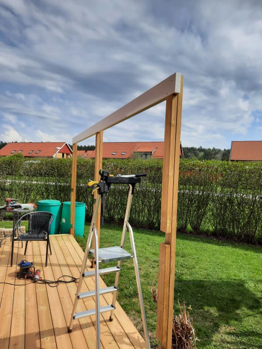 Ny vit bärlina och hammarband på en träaltan under konstruktion, med verktyg och stege i förgrunden och en häck i bakgrunden.