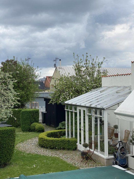 Trädgård med blommande körsbärsäppelträd, växthus och välskött gräsmatta under molnig himmel.