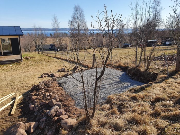 Markarbete med en nyligen breddad kulle, grusväg och markduk i en trädgård med utsikt över en sjö.