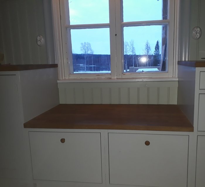 Fönsterplats med bänk och inbyggt skåp i ett ljust rum, med vinterlandskap synligt genom fönstret.
