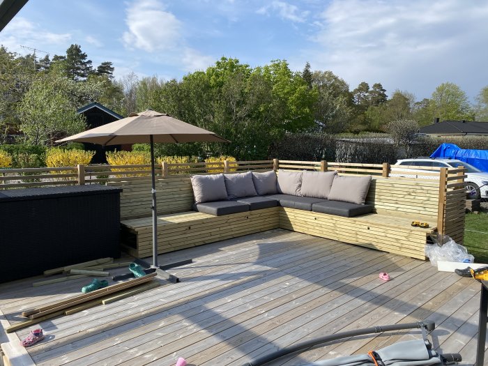 Byggd soffa av trä med dynor på en altan under en parasoll, verktyg och trämaterial synliga.