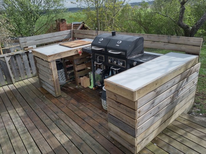 Utekök med gasolgrill och kaklade bänkskivor på trädäck mot en naturbakgrund.