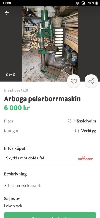 Grön pelarborrmaskin av märket Arboga placerad i ett verktygsrum med ved i bakgrunden.