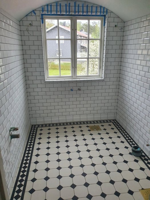 Renoverat badrum med vita väggkakel, mönstrat svart-vitt golvkakel och fönster med utsikt.