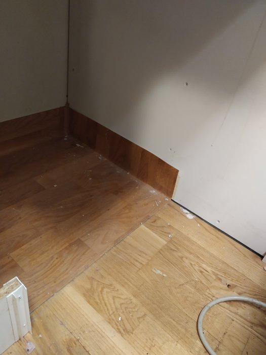 Ett hörn av ett rum med trägolv där delar av en pvc-matta är limmad, visande renoveringsarbete pågår.