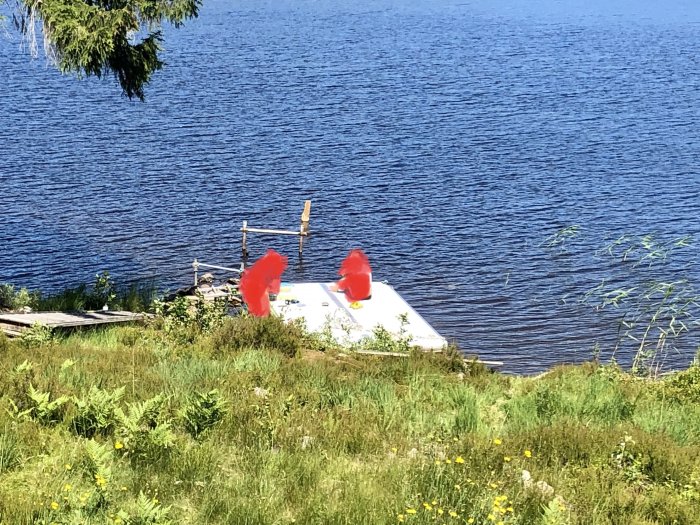 Vy över en renoverad bryggbåt vid en sjö med grön omgivning och röda stolar ombord.