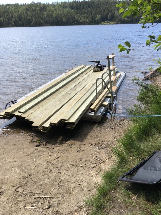 Bryggbåt vid en sjö med nytt virke klart för installation av badstege och fiskespöhållare, i naturlig omgivning.