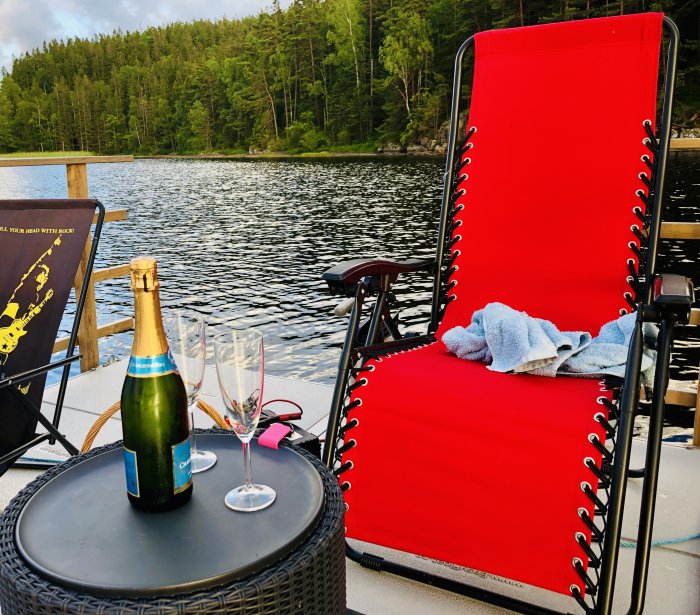 Ett avkopplingsområde på en bryggbåt med en röd solstol, en flaska champagne och tomma glas, med skog och vatten i bakgrunden.