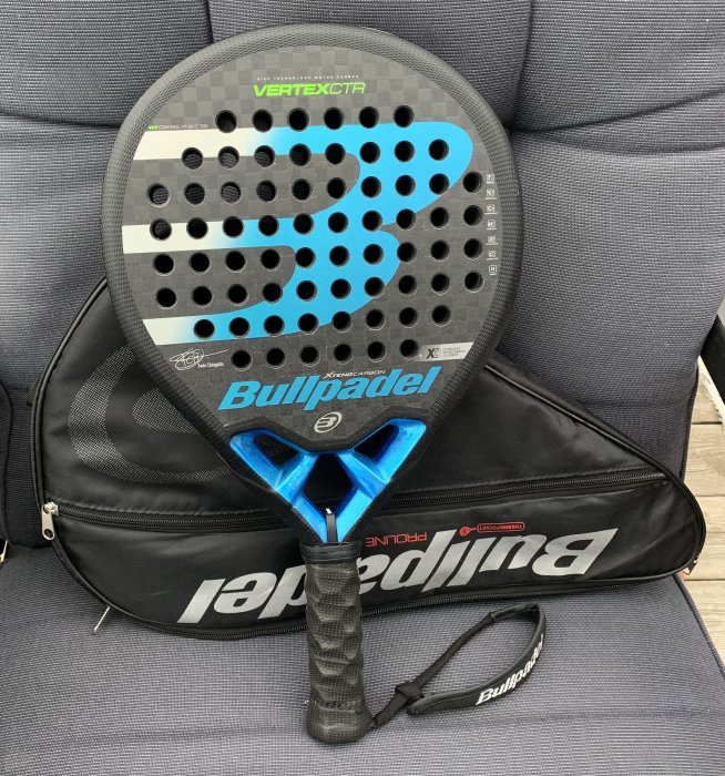 Padelracket från Bullpadel modell Vertex Comfort med hålmönster på stol, ovanpå svart racketväska.