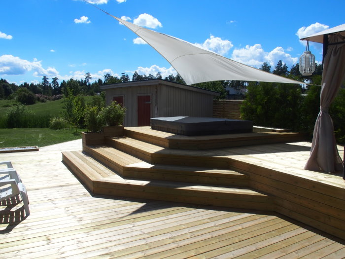 Trädäck i flera nivåer med inbyggda blomlådor och ett solsegel ovanför, bredvid en jacuzzi och ett trädgårdspaviljong.