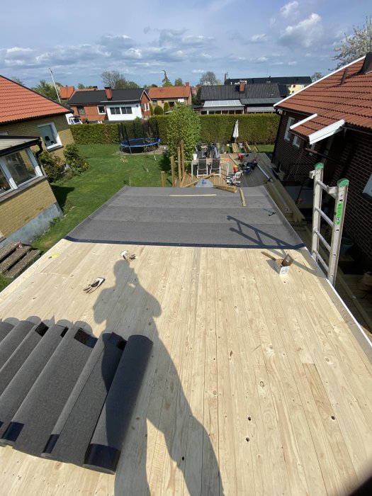 Nyligen utbytt träkonstruktion för garagetak med påbörjad takpappsläggning, verktyg syns, omgivande trädgårdar i bakgrunden.