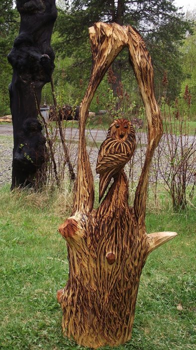 Träskulptur av en uggla uthuggen i en stor stubbe med detaljerade fjädermönster och genombrutna vingar.