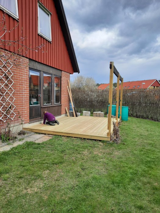En person på knä monterar golvplankor på en altan nära ett rött hus, med nya bärlinor och omonterade takstolar i bakgrunden.