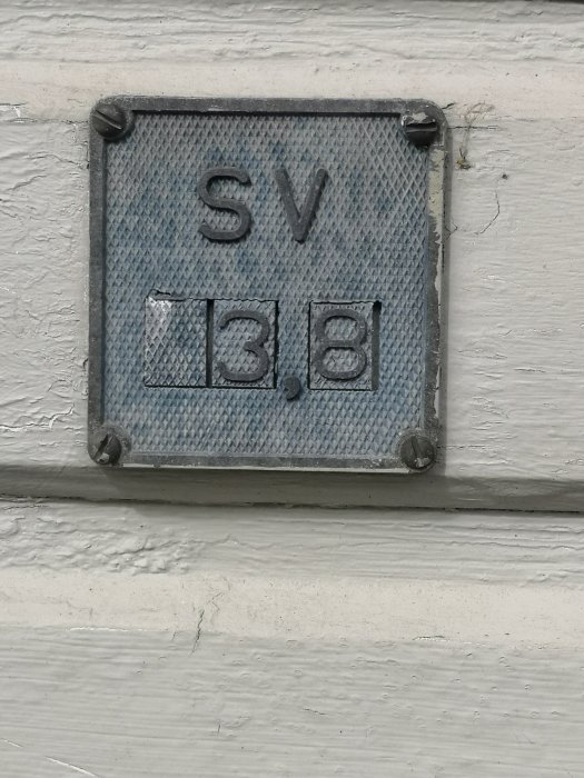 En metallskylt med texten "SV" över siffrorna "13.8" monterad på en vitmålad fasad.