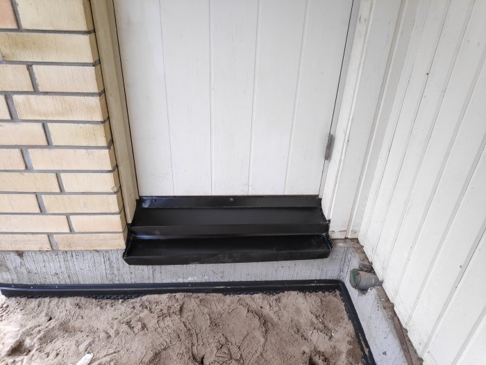Svart tröskelbleck vid dörr, behöver foga, sandig mark, husvägg i bakgrunden