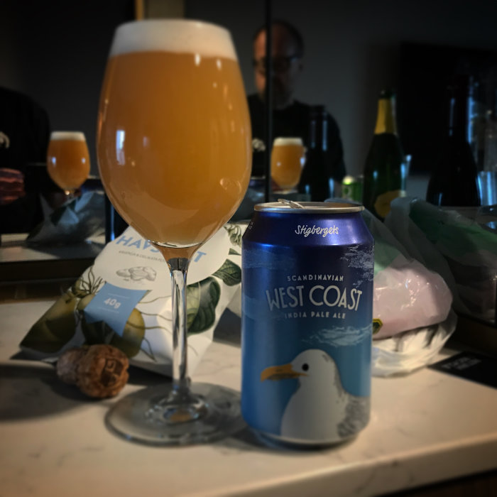 Ölglas fyllt med grumlig öl bredvid burk av West Coast IPA på bänk.