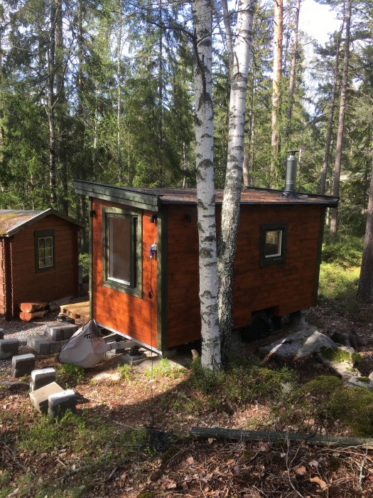 Bastubyggnad med träpanel och metallskorsten i en skogsmiljö, byggmaterial synligt framför, klar för invändig finslipning.