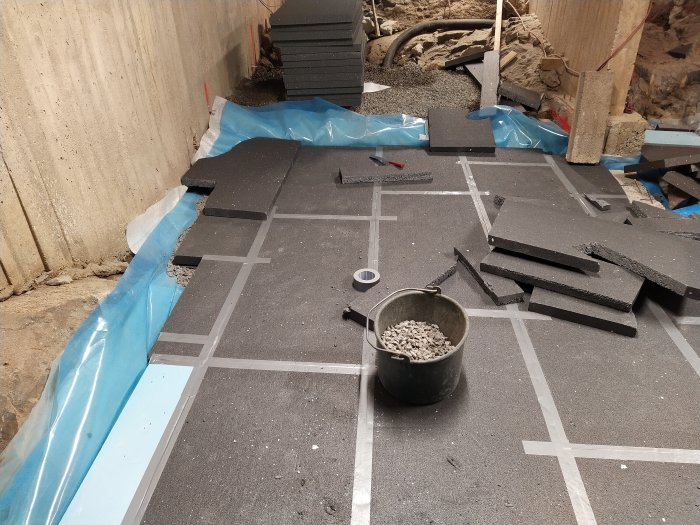 Avjämningsarbete på golv med isoleringsskivor, avskurna bitar och grushink i en byggarbetsmiljö.