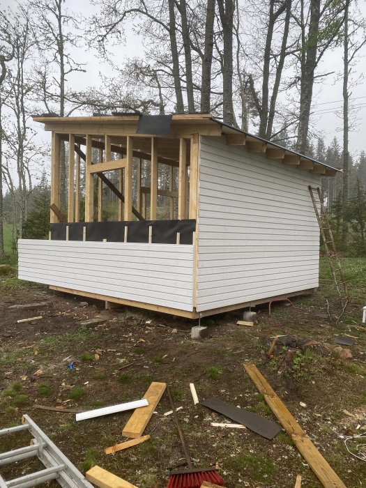 Nypanelad gäststuga under konstruktion med skog i bakgrunden och byggmaterial på marken.