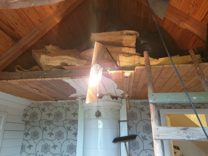 Renovering av tak med öppen isolering och träbjälkar, stege och gammaldags kakelugn i rum.