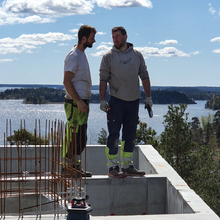 Två personer står på en husgrund i byggkläder med en sjö och skog i bakgrunden.