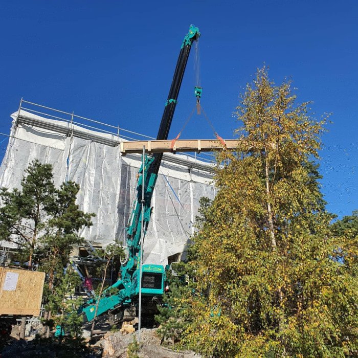 Bygge av egenritat hus på berg med mobilkran som lyfter byggmaterial, omgivet av natur.