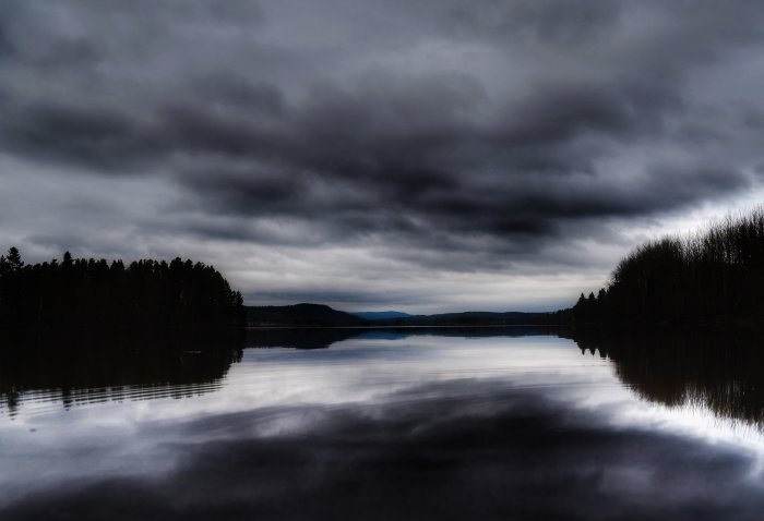 Mörka moln speglas i lugnt vatten med skogklädda kuster på båda sidor.