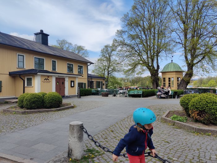 Gatumiljö i Stockholm med ett gult hus och uteservering, gröna träd och barn i förgrunden.
