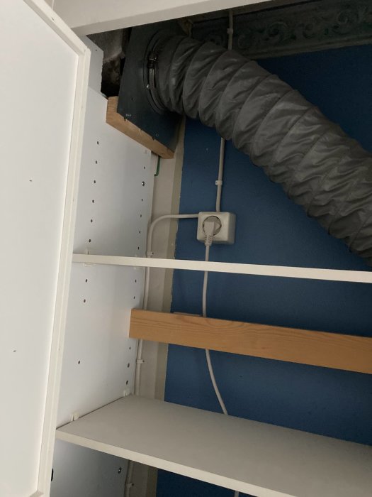 Elektrisk kabel ansluten till ett uttag bakom en tom hylla i ett skåp med synlig ventilationsslang.