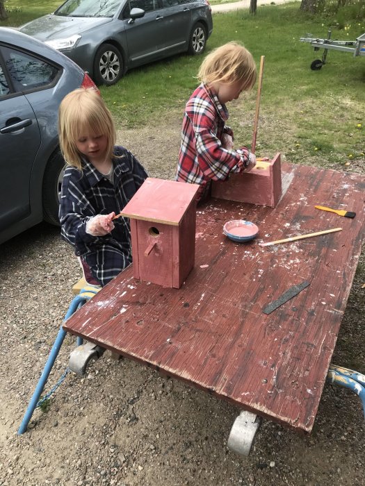 Två barn målar fågelholkar i rosa på ett utomhusbord.