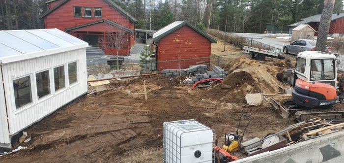 Pågående anläggning av en damm utan fisk, med grävmaskin och byggmaterial runt en grävd grop i en trädgård.