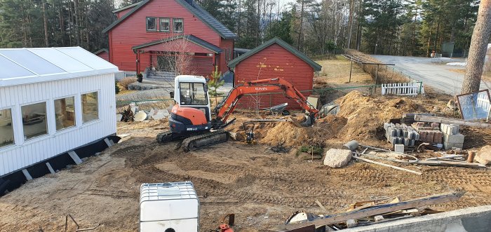 En grävmaskin på en grävd arbetsplats framför ett rött hus, där byggandet av en damm har påbörjats.