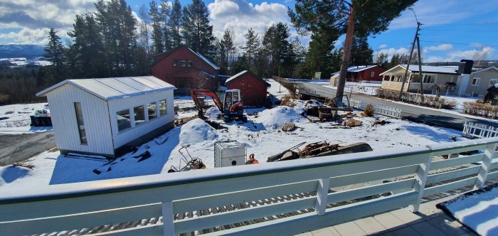 Pågående dammbygge i en snötäckt trädgård med en grävmaskin och byggmaterial runtomkring.