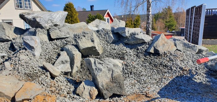 Pågående dammbygge med stora stenblock och grus i en trädgård, ingen vatten eller växter ännu synliga.