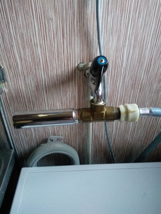 En vattenkran med ansluten genomskinlig tryckdämpare ovanför en tvättmaskin mot en vägg med randig tapet.