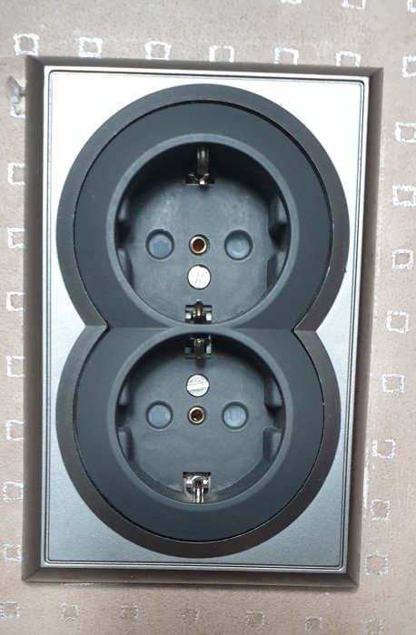 Dubbel vägguttag i svart och silver, europeisk modell monterad på en vägg med tapet.