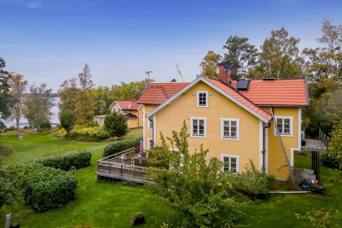 Gult hus med rött sadeltak, grönskande trädgård och blå himmel, potentiellt innan isolering av taket.